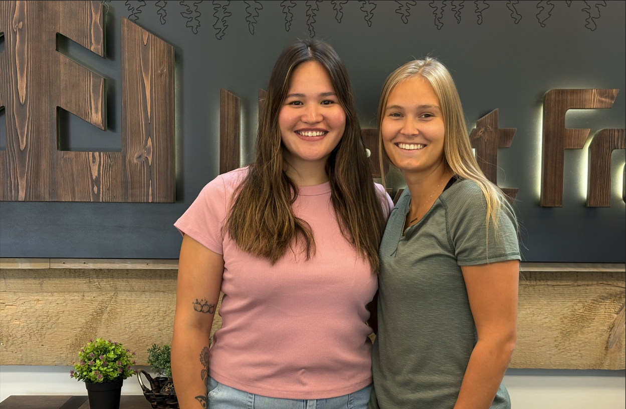 Two women stand against a grey wall with a wooden West Fraser logo.
