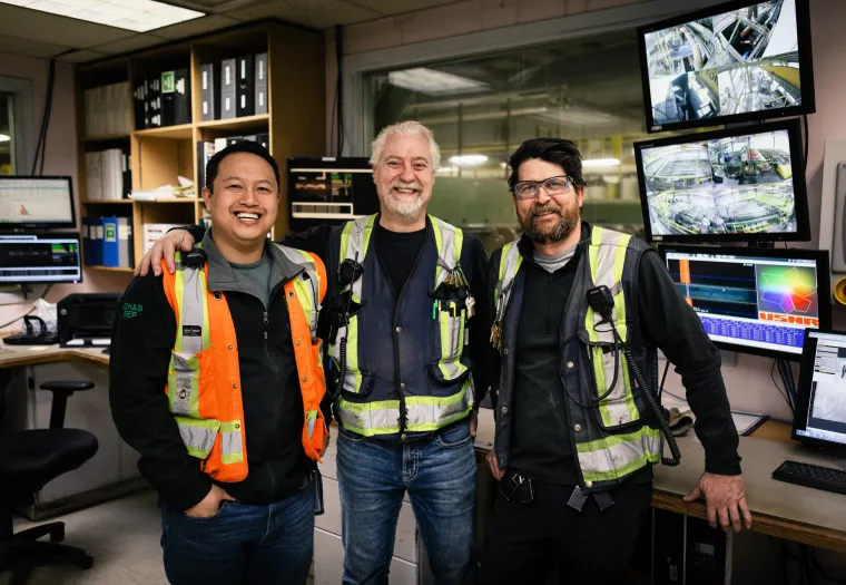 Three men wearing safety vests stand together and smile in a control room with computer monitors and equipment in the background.