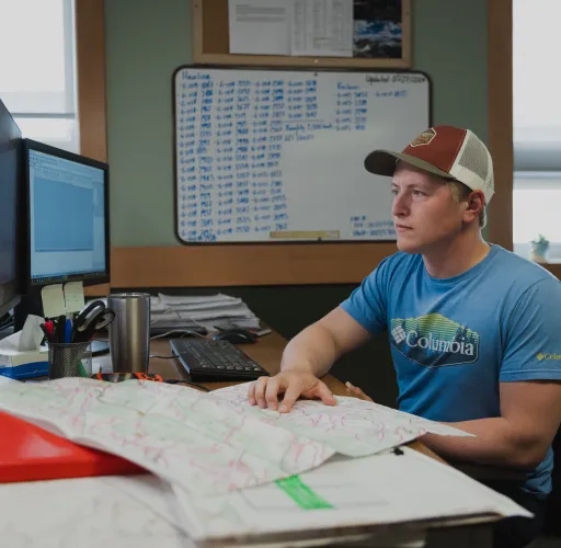 A West Fraser worker wearing a cap and a blue shirt sits at a desk with a computer, keyboard, and maps, surrounded by office materials and a whiteboard in the background.