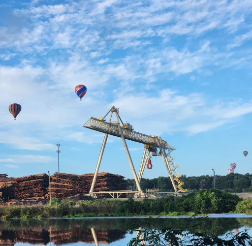 A large industrial crane is stationed near stacks of logs by a water body. Several hot air balloons are floating in the sky above, against a backdrop of trees and a clear blue sky.