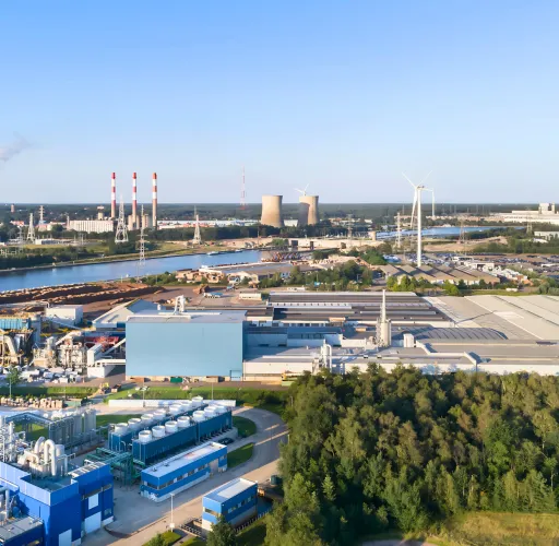 Aerial view of an industrial complex with factories, smokestacks emitting smoke, warehouses, wind turbines, and surrounding greenery under a clear sky.