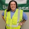A person wearing a yellow safety vest stands smiling in front of a green and white banner.